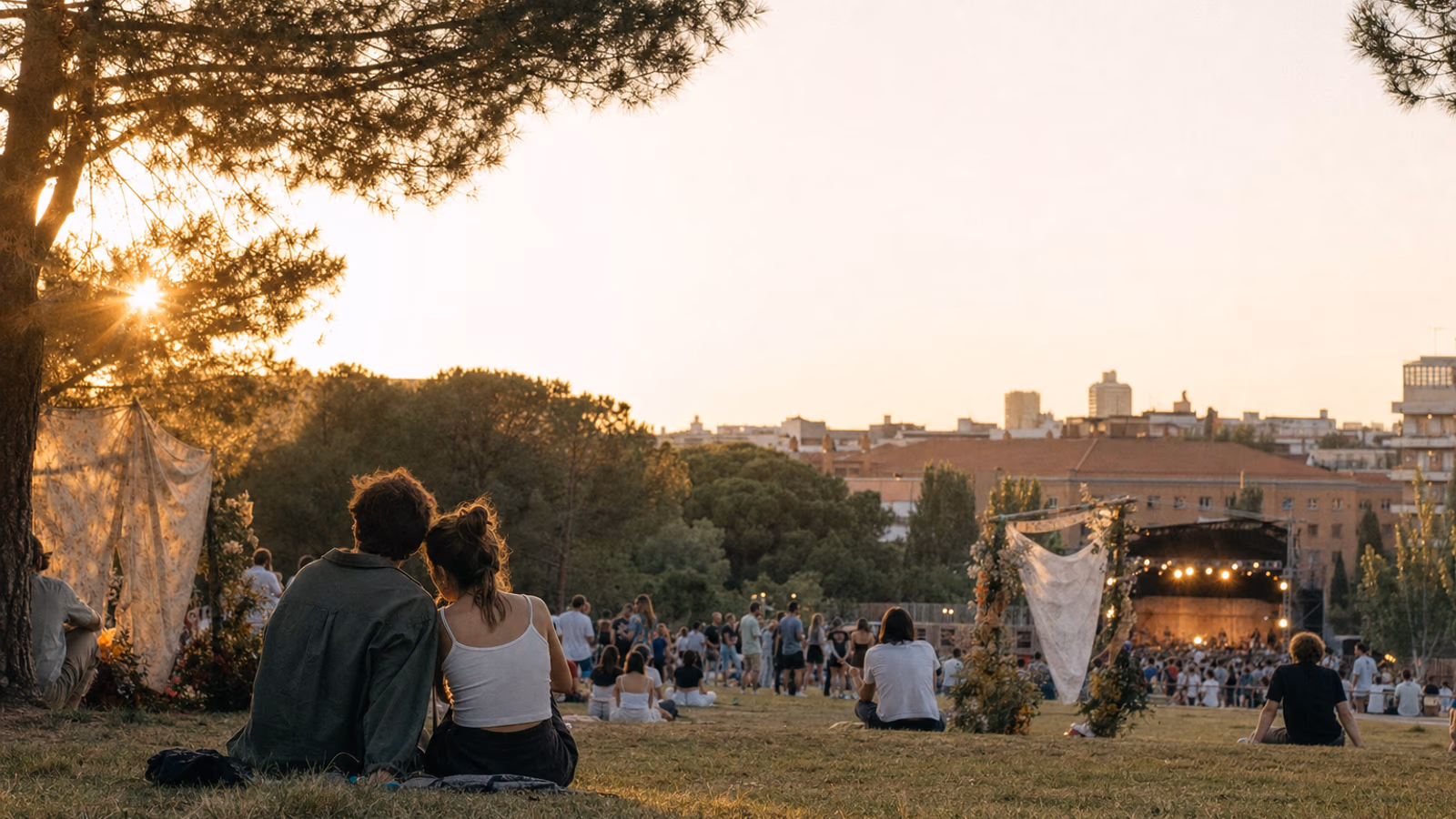 Festival-goers sitting on the grass at sunset facing a stage among pine trees